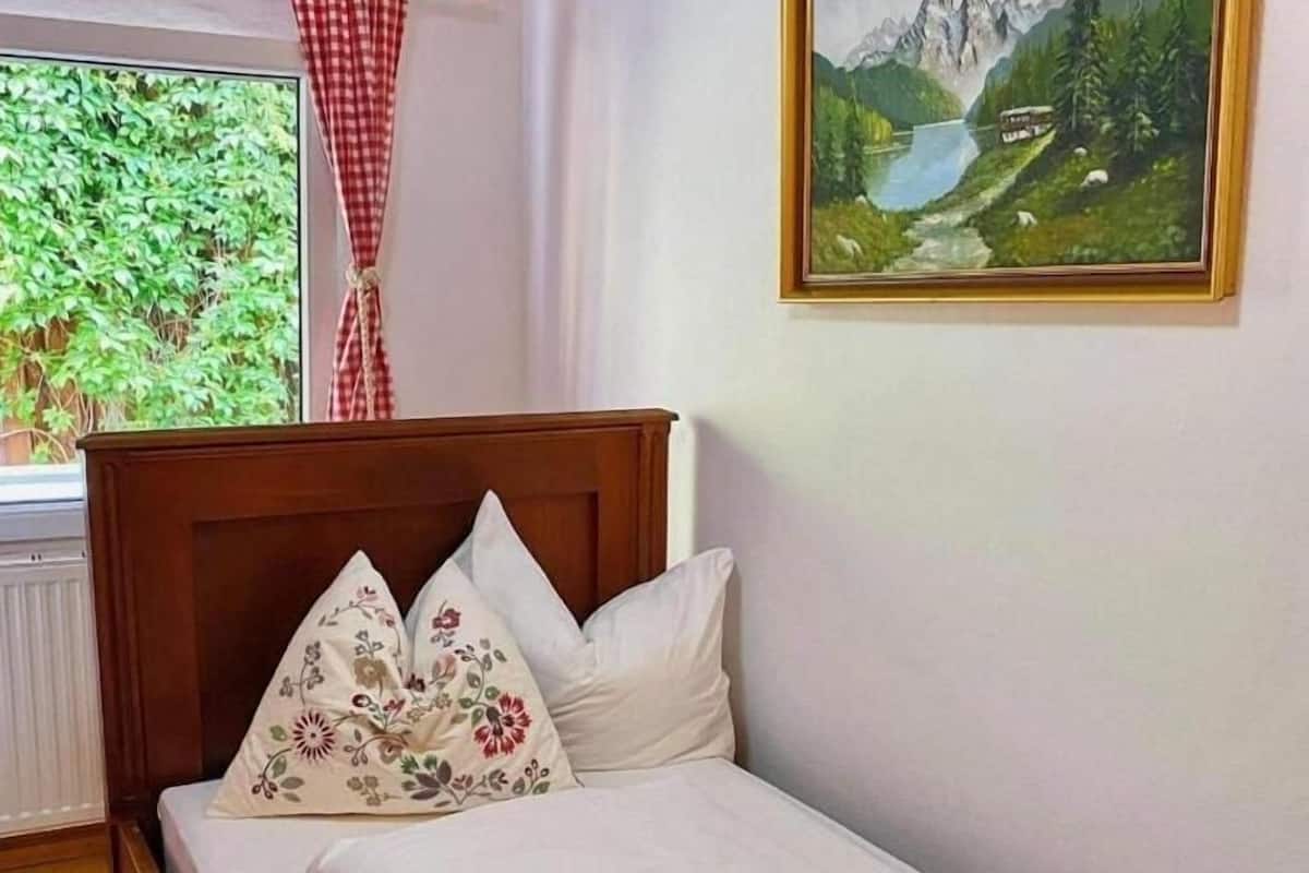 Een slaapkamer met een houten bed, een raam met uitzicht op groen, en een schilderij van een berglandschap aan de muur.