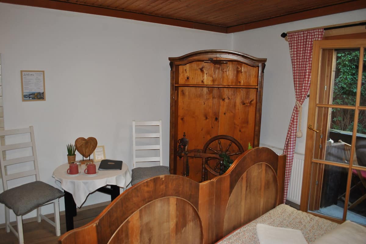 Een kamer met een houten bed, een klein tafeltje met een laptop, een stoel, een kledingkast en een raam met een geruit gordijn.