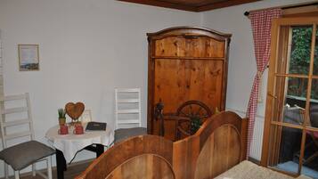 A room with a wooden bed, a small table with a laptop, a chair, a wardrobe, and a window with a checkered curtain.