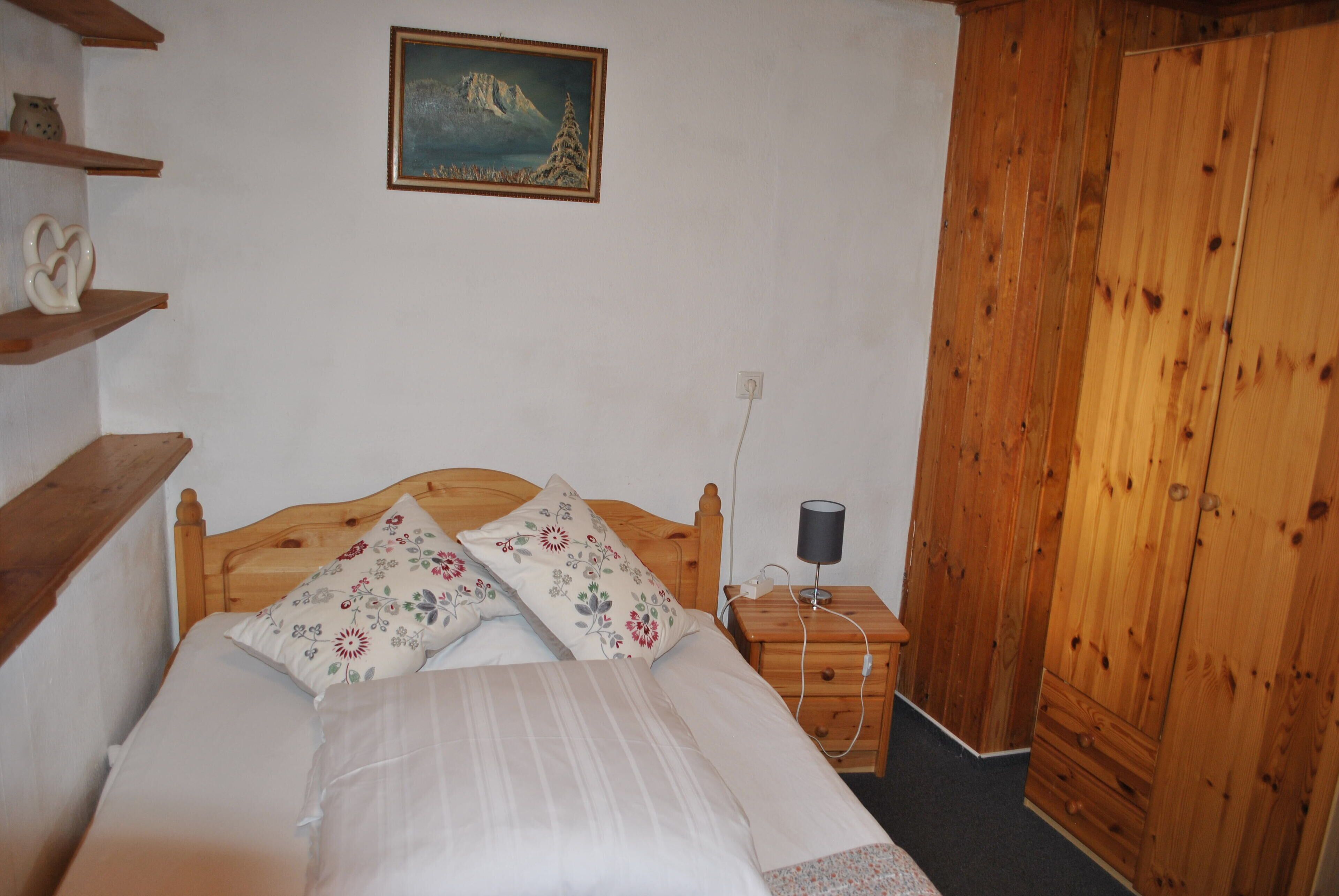 Un dormitorio con cama de madera, mesita de noche con lámpara, un cuadro en la pared y un armario de madera.