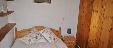 A bedroom with a wooden bed, a nightstand with a lamp, a wall-mounted painting, and a wooden wardrobe.