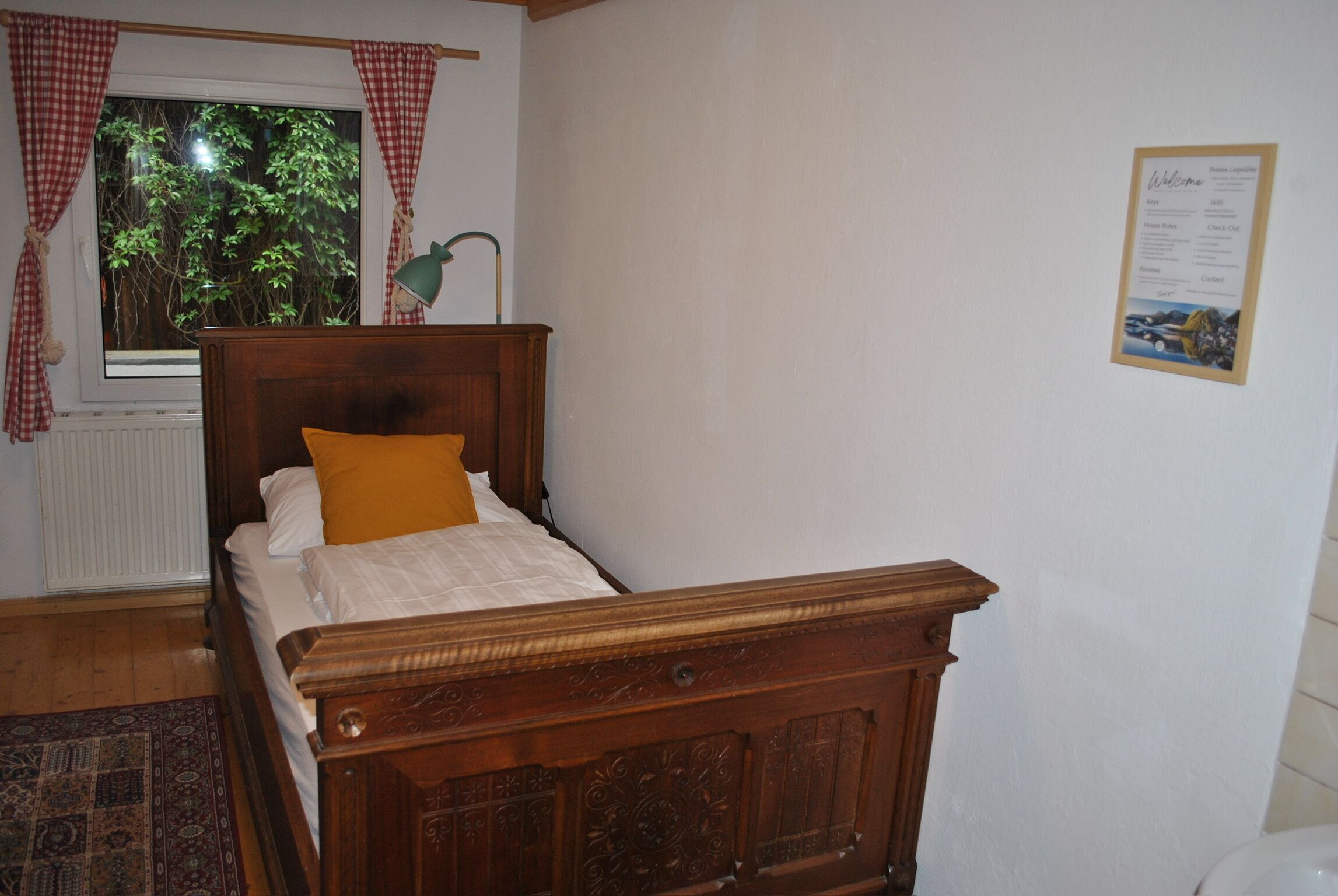 Una cama de madera con un almohadón blanco y amarillo, una ventana con cortinas rojas y blancas, y un cartel de bienvenida en la pared.