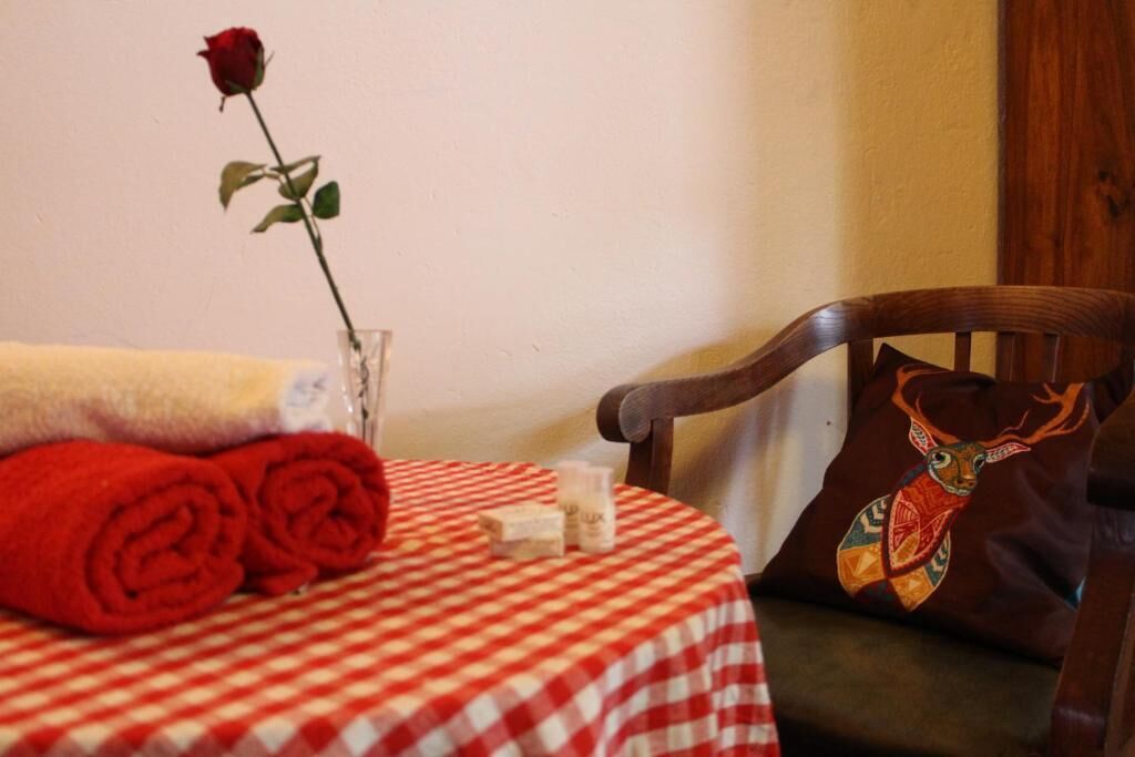 Una mesa redonda con mantel a cuadros, un florero con una sola rosa, toallas enrolladas y una silla con un cojín decorativo.
