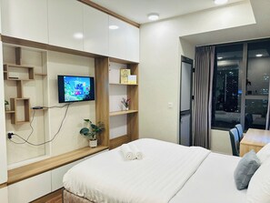 Una habitación de hotel con una cama, un televisor fijado a la pared, una planta pequeña y una ventana con cortinas.