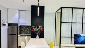 A modern kitchen with a central island, a dining area with chairs, a TV, and a decorative vase with flowers. - SS Homes River Gate Premium (Ho Chi Minh City)
