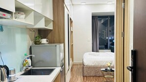 A compact hotel room with a kitchenette, a bed, a small table with a vase, and a view of the city at night. - SS Homes River Gate Premium (Ho Chi Minh City)