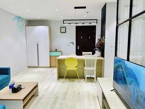 A modern living room with a sofa, a dining table with chairs, a TV, and a kitchen area with a fridge and a sink. - SS Homes River Gate Premium (Ho Chi Minh)