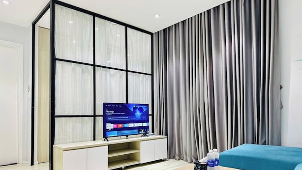 A modern living room with a flat-screen TV on a white entertainment unit, a blue sofa, and a wooden coffee table with a remote control and water bottles. - SS Homes River Gate Premium (Ho Chi Minh City)
