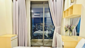 A hotel room with a bed, a nightstand, a dresser, a mirror, and a view of a cityscape at night. - SS Homes River Gate Premium (Ho Chi Minh City)