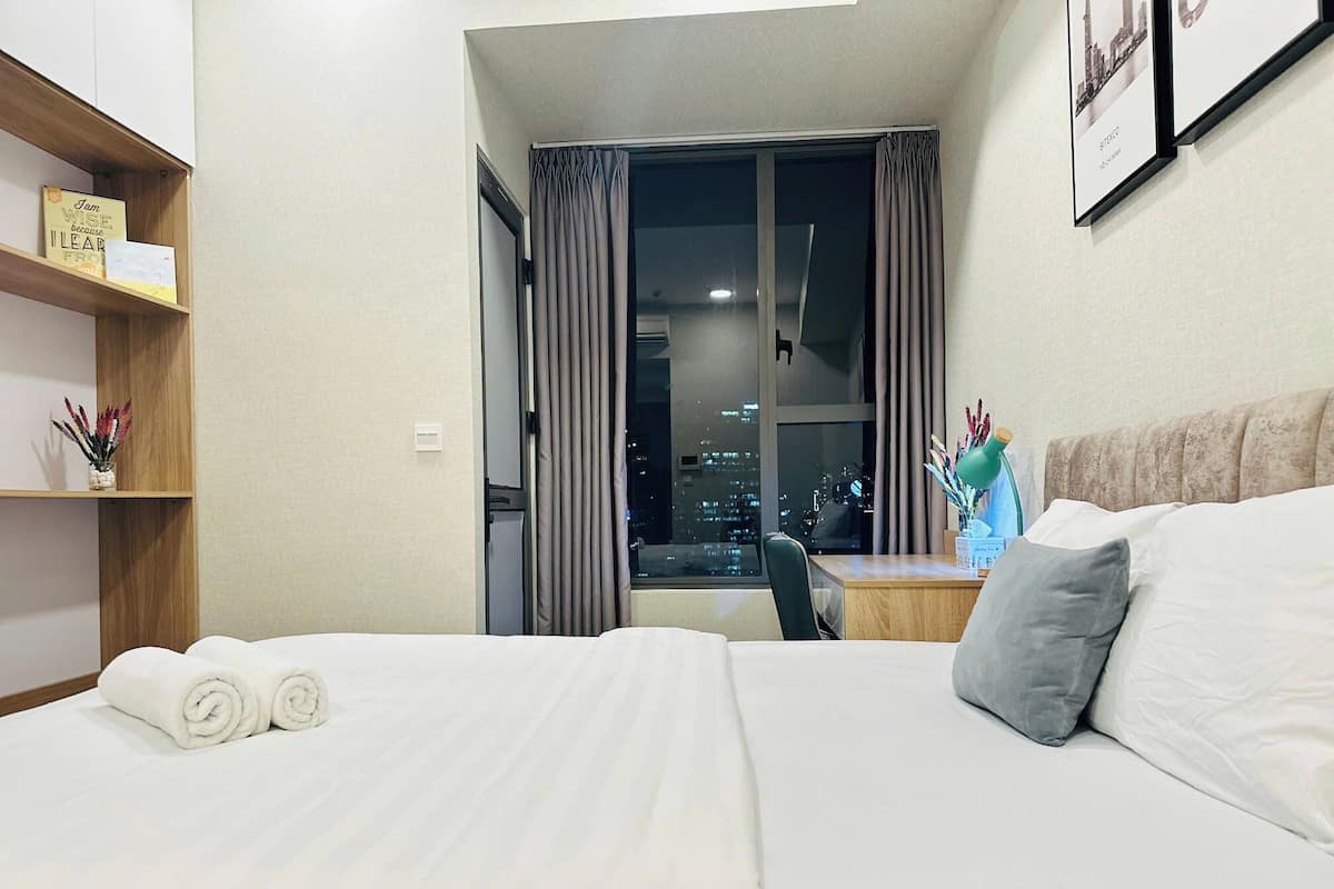 Ein Hotelzimmer mit Bett, Schreibtisch, Stuhl, einem Fenster mit Vorhängen und einem Regal mit Büchern und einer Pflanze.