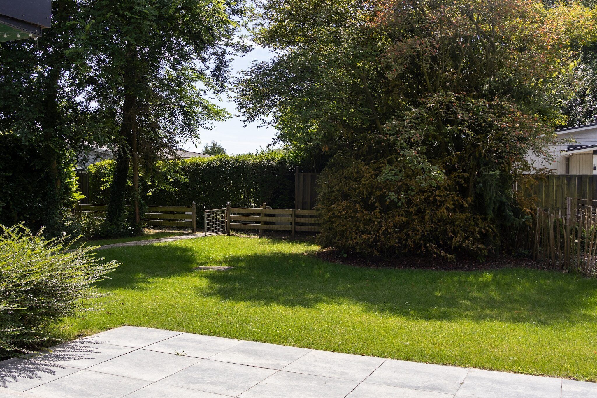 A well-maintained garden with a wooden fence, a paved walkway, and various shrubs and trees.