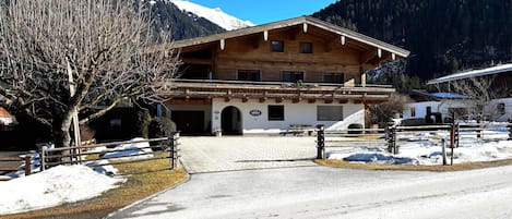 A wooden chalet with a balcony, a paved driveway, and a mountain backdrop.