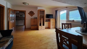 A room with a wooden floor, a dining table with chairs, a television, a cabinet, and a window with curtains. - Apartment in a quiet location with beautiful view (Neukirchen am Großvenediger)