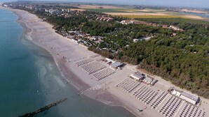 Vista aérea de una playa con filas de sombrillas y sillas de playa, un estacionamiento y una zona residencial con casas y vegetación al fondo.
