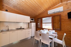 A compact kitchen with white cabinets, a microwave, a refrigerator, and a dining area with white chairs and a table. - Bungalow in the Adriano Family Camping Village (Punta Marina)