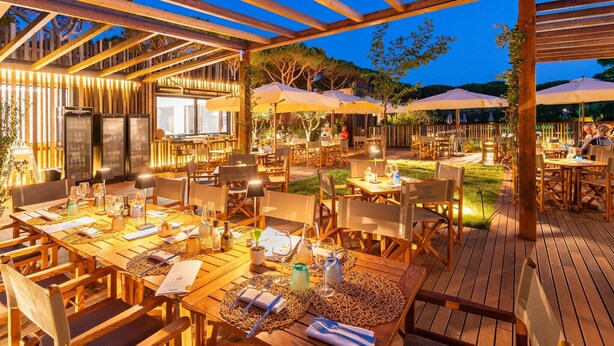 Un área de comedor al aire libre con mesas y sillas de madera, sombrillas y una estructura de pérgola.