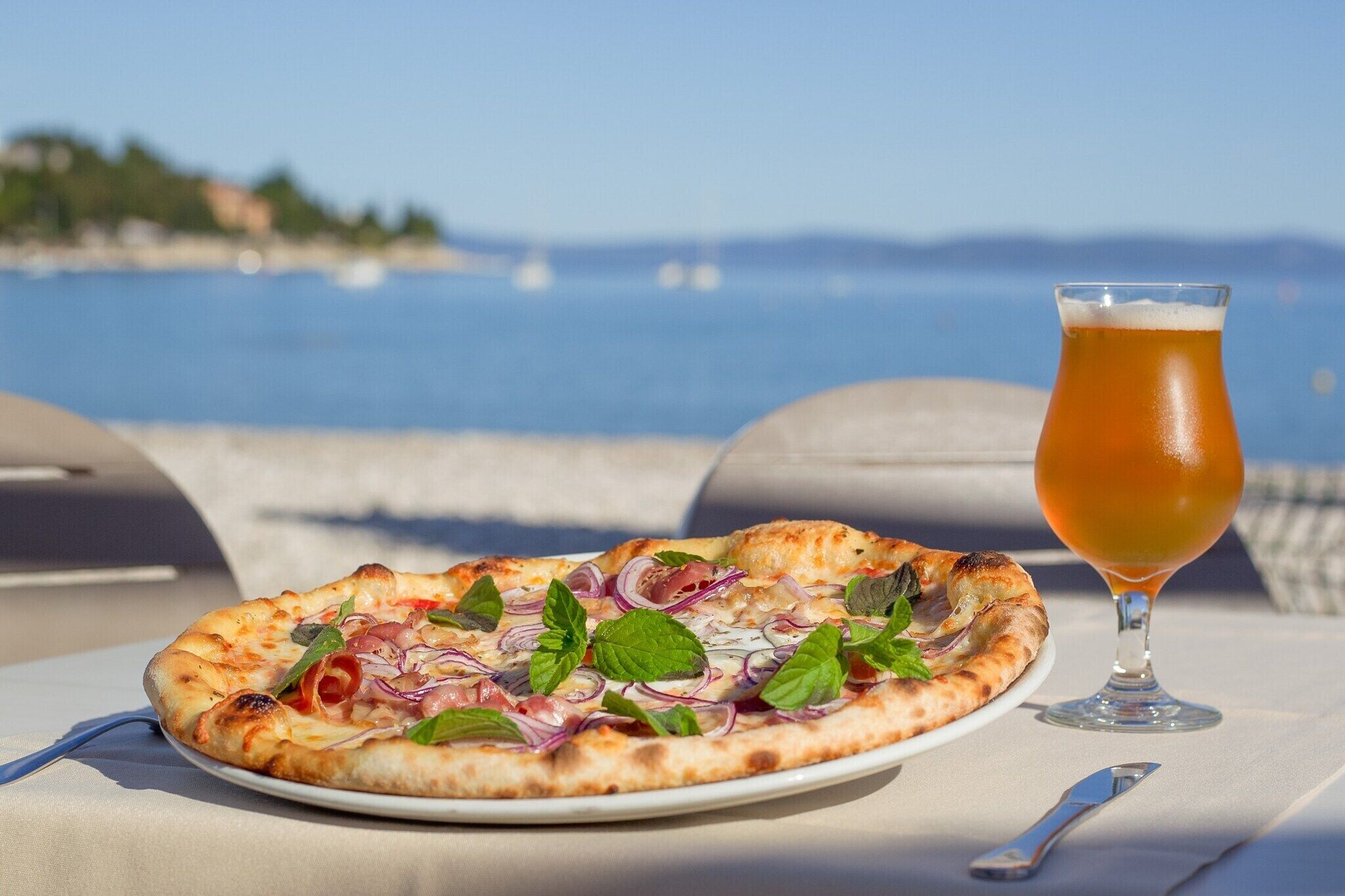 A pizza with toppings and a glass of beer on a table by the sea.