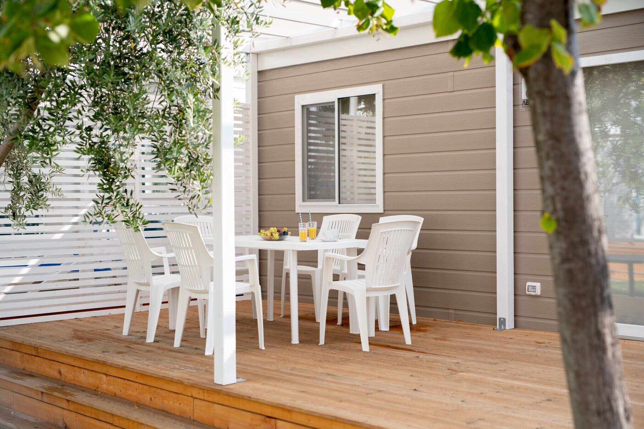 Un patio cubierto con una mesa y sillas blancas, una ventana con persianas y una terraza de madera.