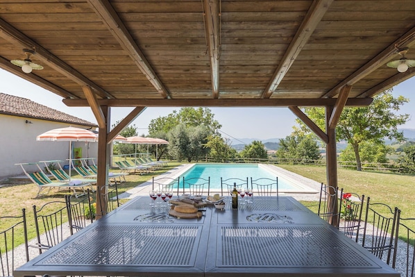 A covered outdoor area with a pool, lounge chairs, umbrellas, and a table set for dining.