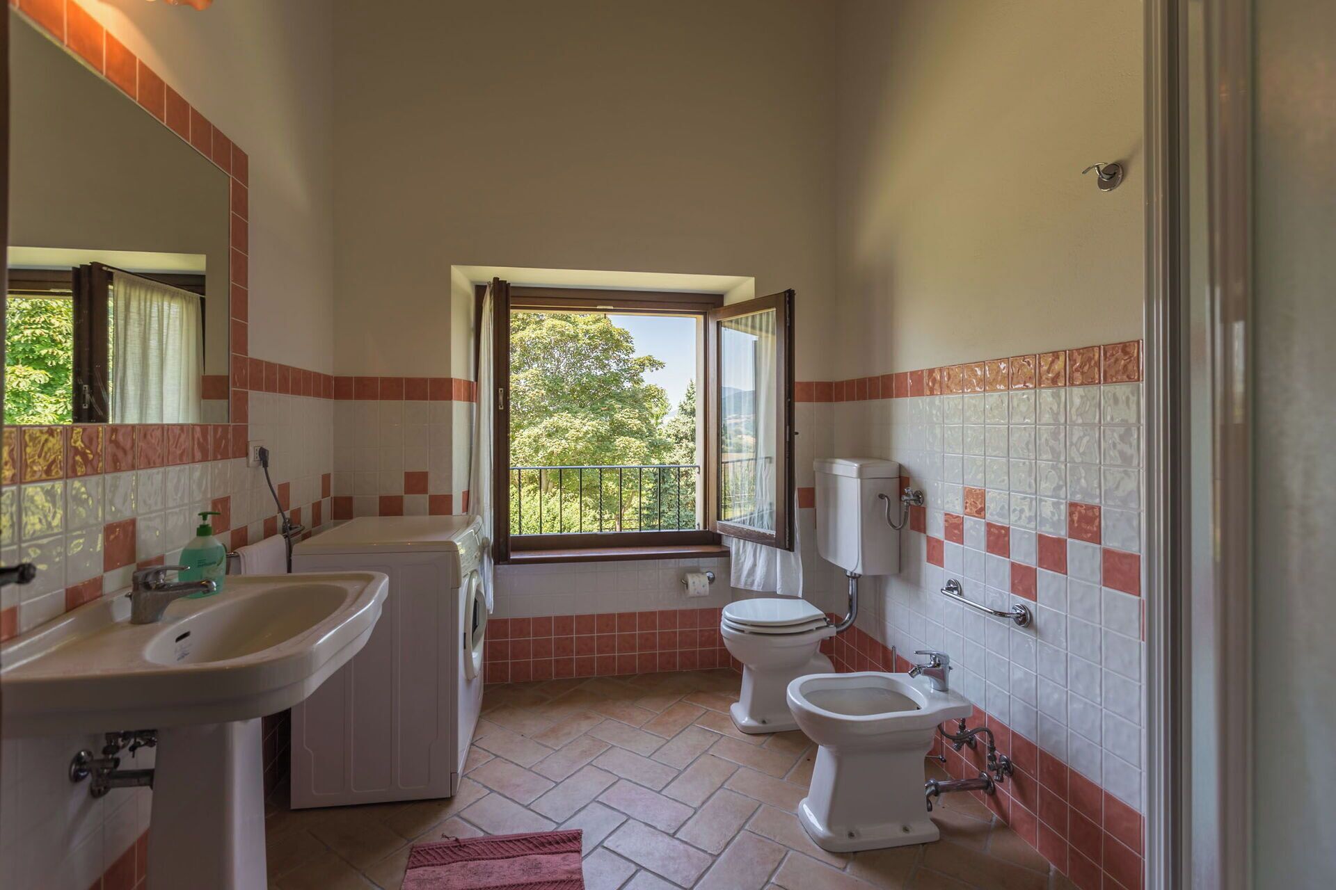 Casa de banho com lavatório, duas sanitas, máquina de lavar roupa e janela com vista para árvores.