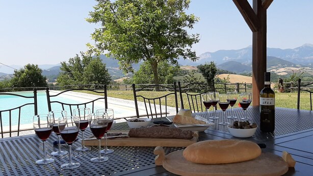 Ein Tisch ist für eine Mahlzeit gedeckt, mit Weingläsern, einer Weinflasche, Brot und einer Oliven-Schale, mit Blick auf einen Pool und Berge.