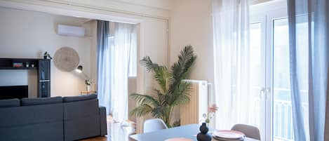 A modern living room with a sofa, dining table, chairs, and a large window with curtains.
