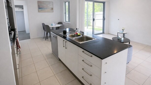 Una cucina moderna con piano di lavoro nero, lavello in acciaio inossidabile e mobili bianchi. C'è un'area pranzo con tavolo e sedie, e una finestra che offre luce naturale.
