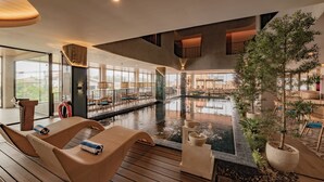 Un espace piscine intérieur moderne, doté de chaises longues, d’un élément décoratif aquatique et offrant une vue sur l’extérieur.