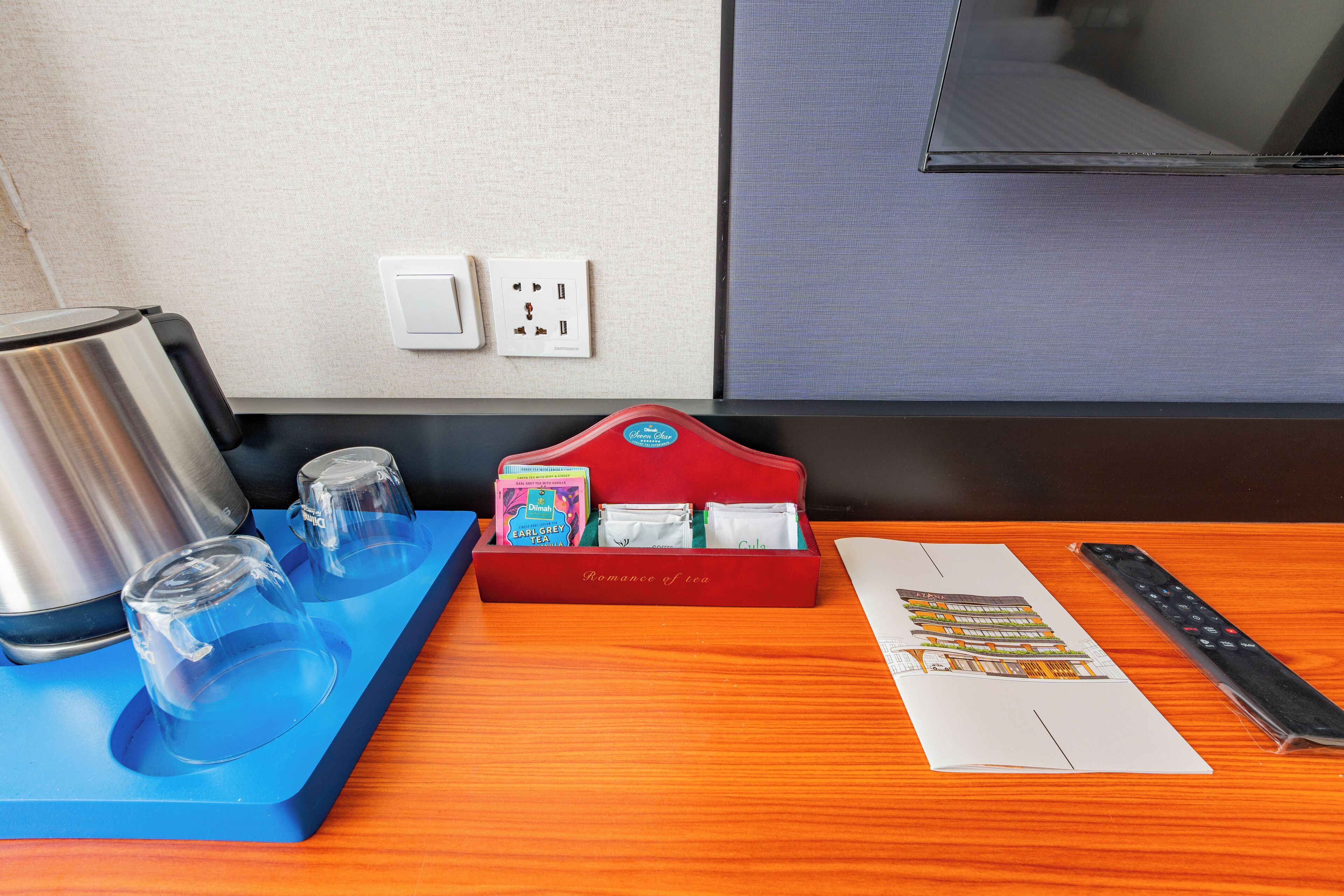 A hotel room with a wooden table, a red teapot set, a remote control, and a menu on the table.