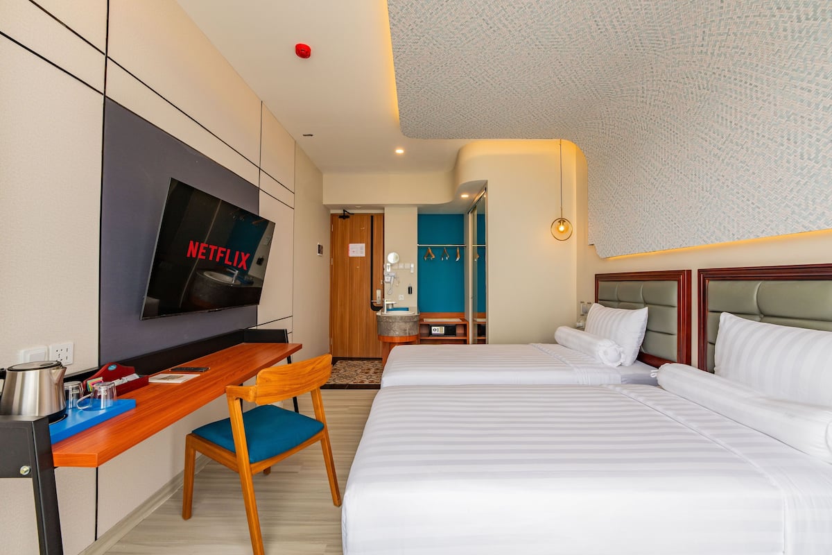 Une chambre d’hôtel avec deux lits, un bureau avec une chaise, une télévision et une salle de bain visible à travers une porte ouverte.