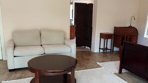 A living room with a beige sofa, a round wooden coffee table, a small wooden side table, a dark wooden cabinet, and a patterned rug. - Peaceful spacious studio in Vista surrounded by greenery and birdsong. (Vista)