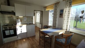 A compact kitchen with a stove, sink, and refrigerator. There is a dining area with a table and chairs. The room has a window with curtains and a view of trees outside. - FeWo Fokko - Away from the hustle and bustle (Norden)