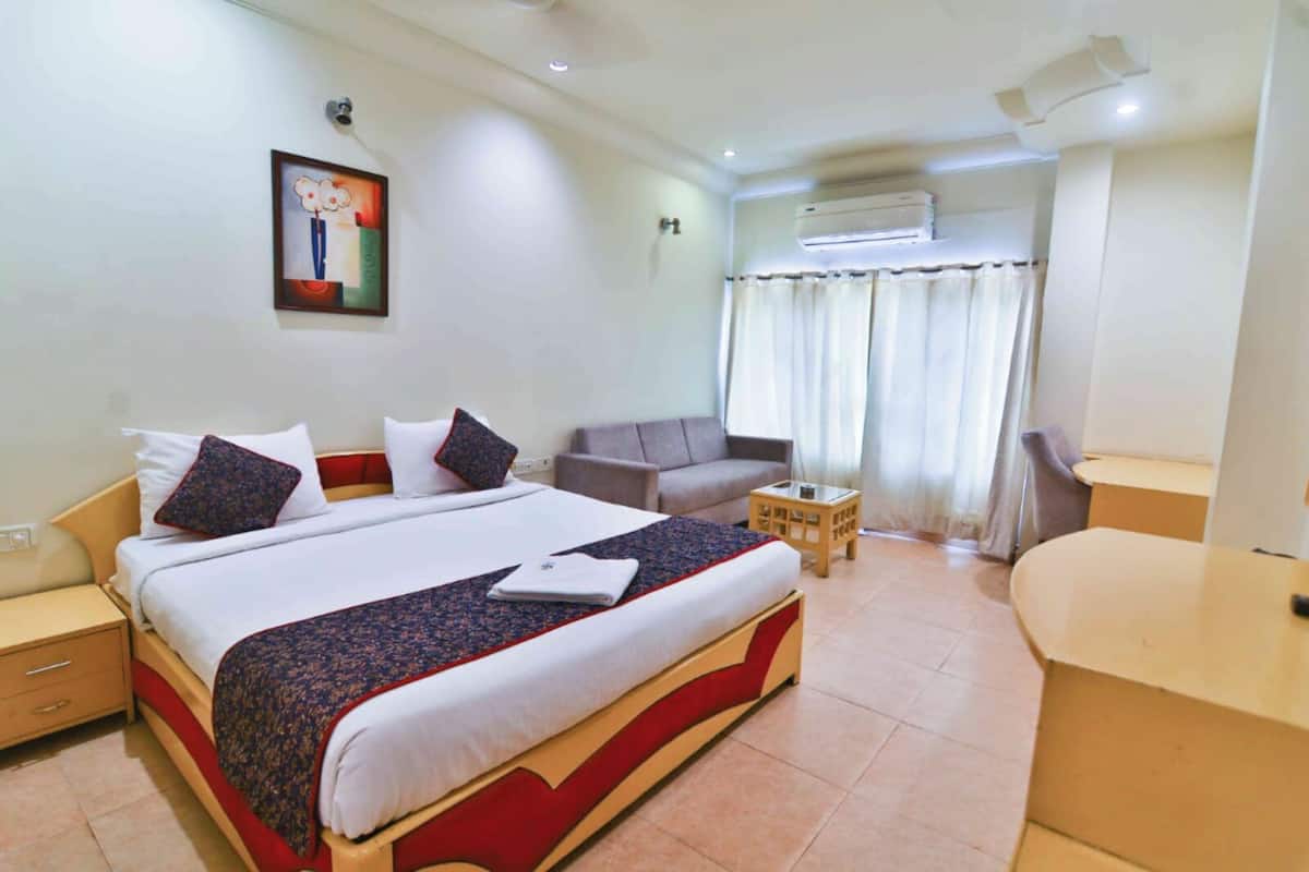 A hotel room with a large bed, a sofa, a small table, and an air conditioning unit.