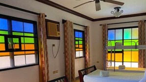 Une chambre avec deux lits, un ventilateur de plafond et des fenêtres offrant une vue colorée.