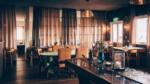 Een restaurant met houten tafels en stoelen, een bar en grote ramen met gordijnen.