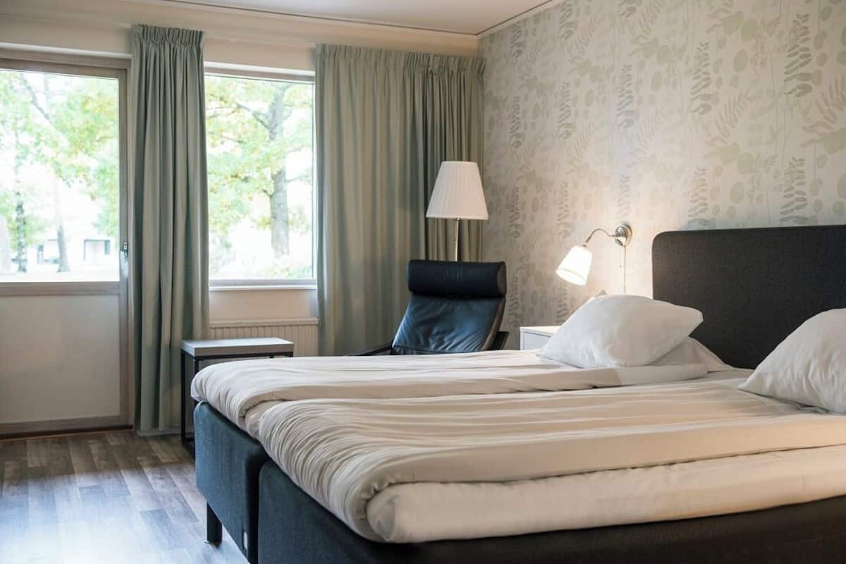 A hotel room with a bed, a chair, a lamp, and a window with curtains.