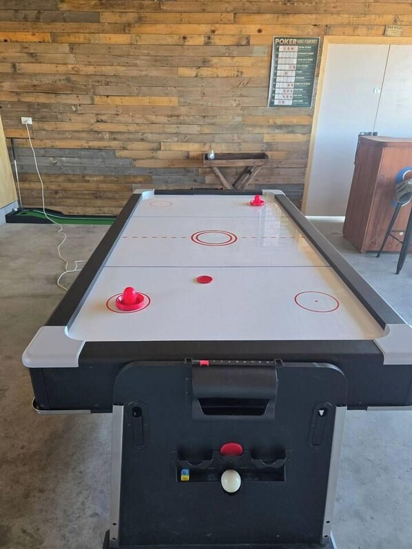 A foosball table with four red and one white ball, set against a wooden wall with a poker table in the background.