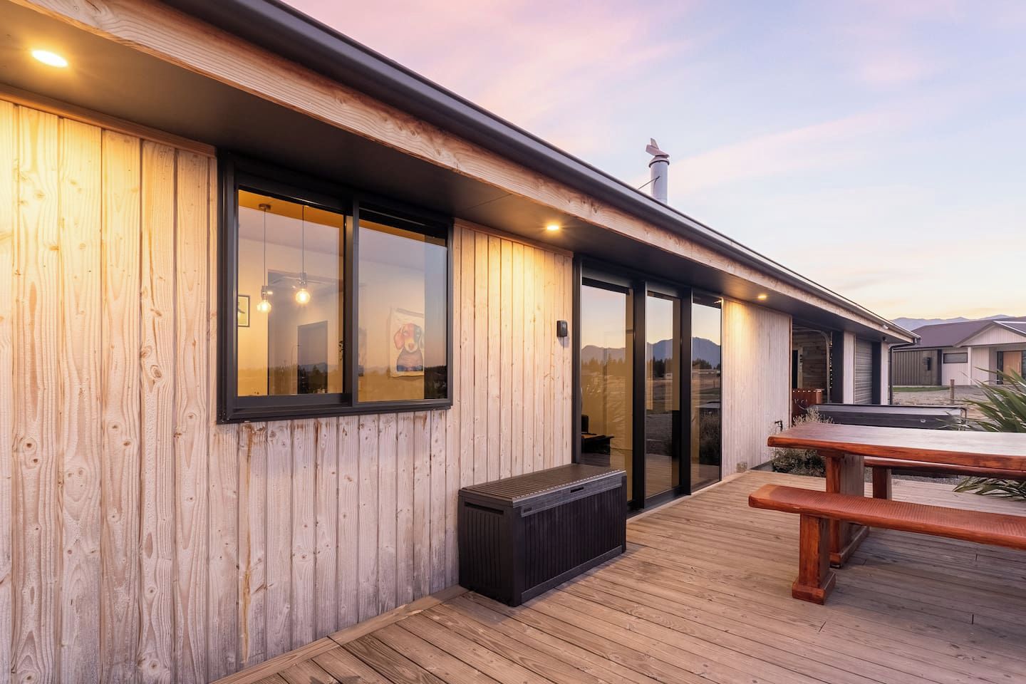 A wooden deck with a bench and a table, a glass door leading to a covered outdoor area, and a view of the mountains in the background.