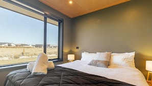 Een slaapkamer met een groot raam, een bed met een donkere quilt en uitzicht op een veld en gebouwen.