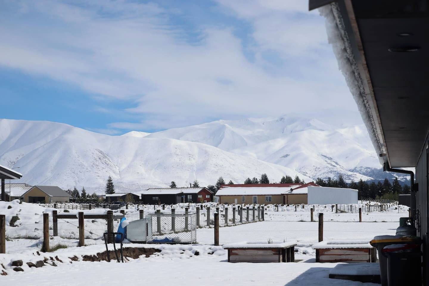 A snowy landscape with a mountain in the background, a fenced area with benches, and buildings with snow-covered roofs.
