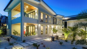 Una casa moderna de dos pisos con un balcón, un jardín bien iluminado y cielo despejado.