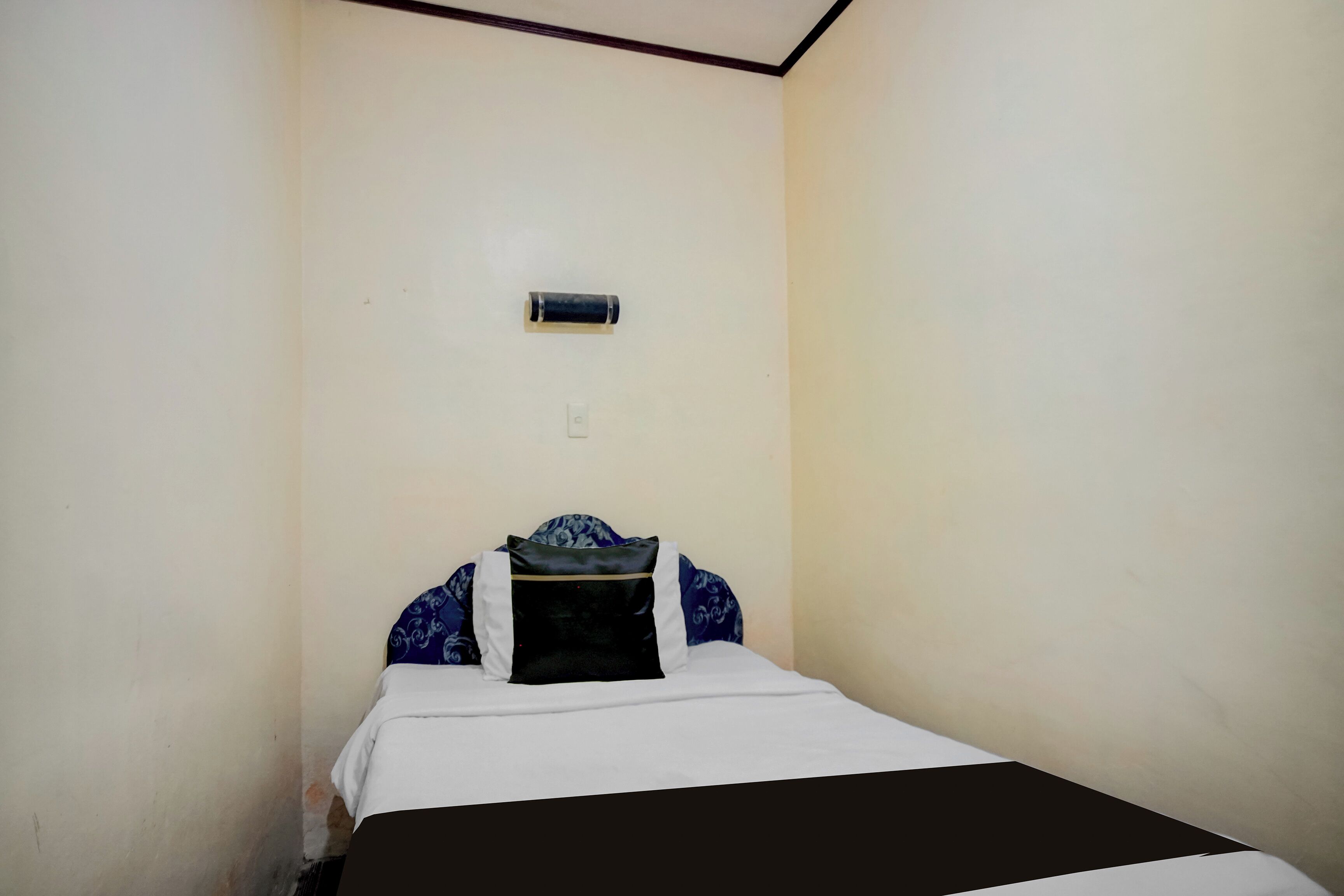A small, single-bed room with a plain wall, a small air conditioning unit, and a bed with a white and black bedspread.