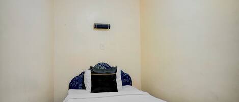 A small, single-bed room with a plain wall, a small air conditioning unit, and a bed with a white and black bedspread.