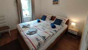 Un letto rifatto con cura, con copriletto a tema nautico, un comodino con una lampada e una finestra con tende.