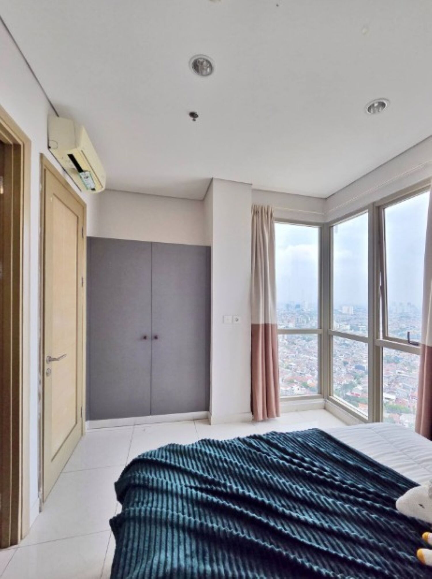 Quarto com uma cama grande, um guarda-roupa e vista para a cidade através de janelas amplas.