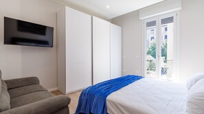 A bedroom with a bed, a TV mounted on the wall, a grey sofa, and a sliding door leading to an outdoor area. - Casa Valentina | WiFi Stazione Treni Parking (Modena)