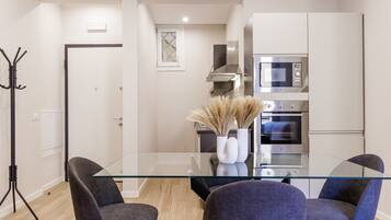 A modern kitchen with a glass dining table, two chairs, a microwave, an oven, and a decorative vase with dried plants.
