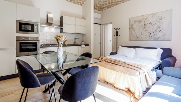 A modern hotel room with a glass dining table, dark upholstered chairs, a bed with a beige duvet, and a large abstract painting on the wall. - House Chiara| WiFi Parking Station downtown Modena (Modena)