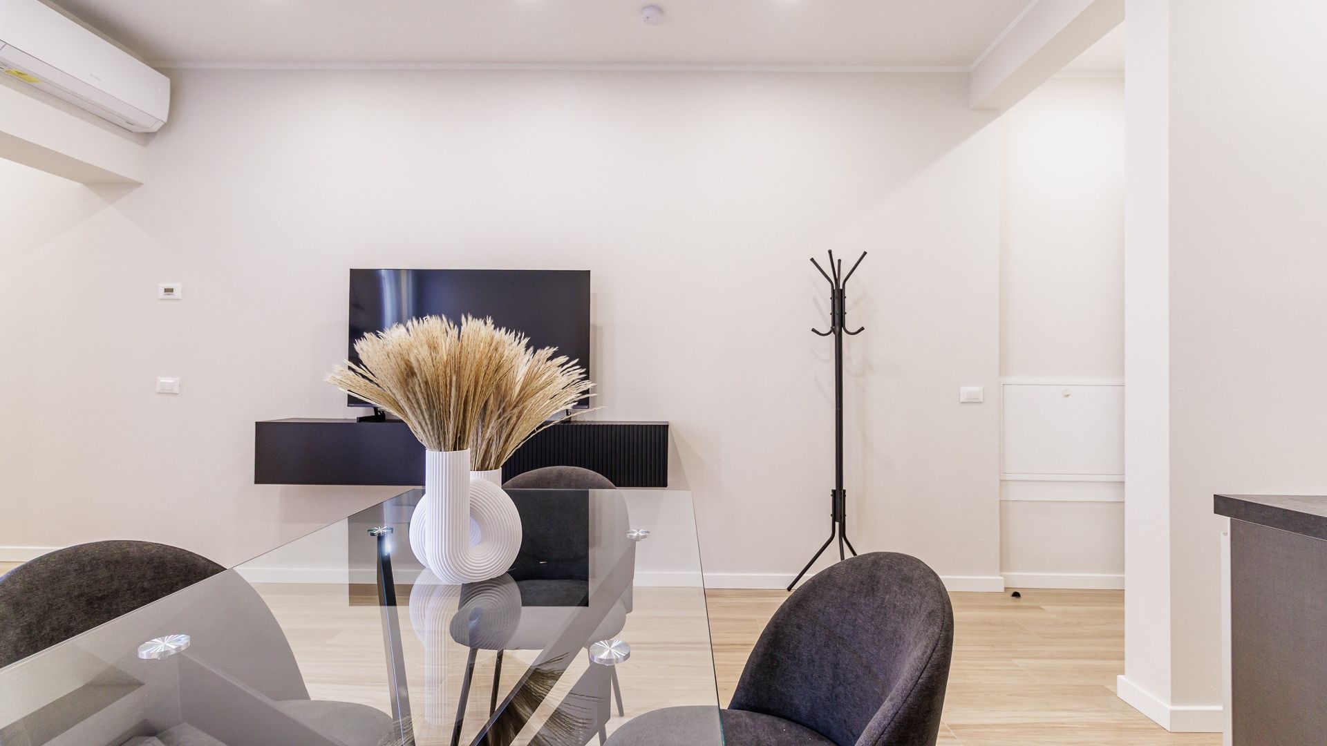 Un salon moderne avec une table à manger en verre, deux chaises, un vase contenant des plantes séchées, un porte-manteau noir et un téléviseur à écran plat fixé au mur.