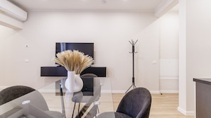 Ein modernes Wohnzimmer mit einem Glastisch, zwei Stühlen, einer Vase mit Trockenpflanzen, einem schwarzen Kleiderständer und einem flachbildfernseher an der Wand.